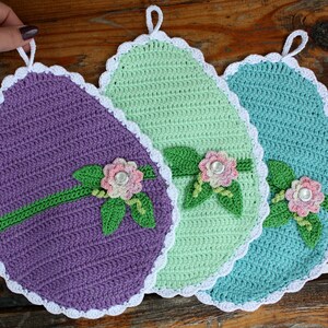 Puede incluir: Tres agarraderas de olla de ganchillo en morado, verde y azul con ribete blanco y detalles florales. Cada agarradera tiene un lazo para colgar.