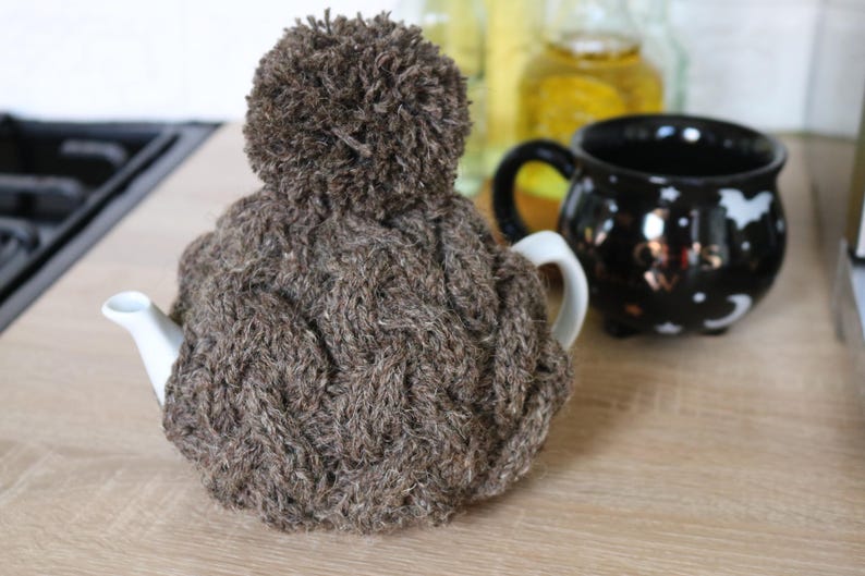May include: A brown knitted teapot cozy with a pom-pom on top. The cozy is covering a white teapot with a spout.