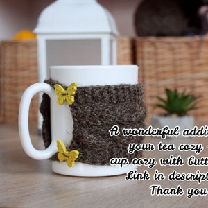 May include: A white ceramic mug with a brown knitted cozy featuring yellow butterfly embellishments. The text "A wonderful addition to your tea cozy - a cup cozy with butterflies. Link in description. Thank you" is printed below the mug.