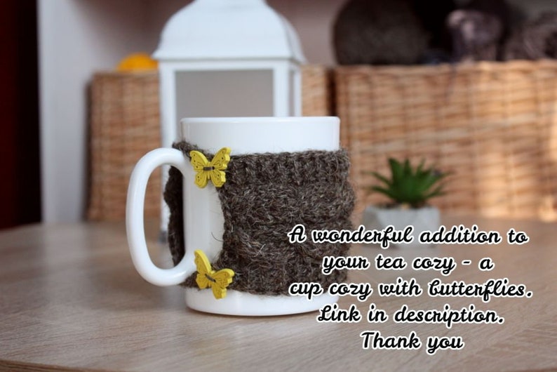 May include: A white ceramic mug with a brown knitted cozy featuring yellow butterfly embellishments. The text "A wonderful addition to your tea cozy - a cup cozy with butterflies. Link in description. Thank you" is printed below the mug.
