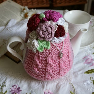 May include: A pink knitted teapot cosy with crocheted flowers in shades of white, pink, and burgundy. The cosy is decorated with a silver charm.