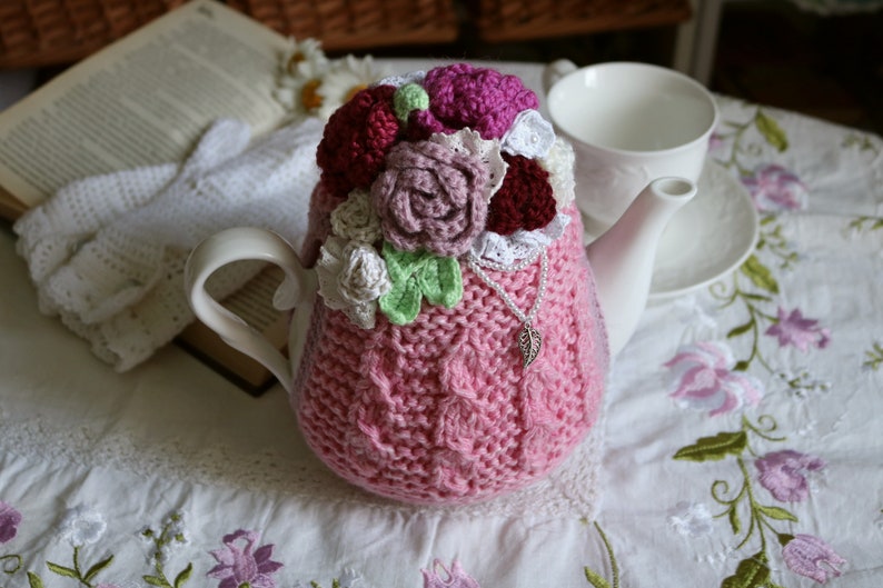 May include: A pink knitted teapot cosy with crocheted flowers in shades of white, pink, and burgundy. The cosy is decorated with a silver charm.
