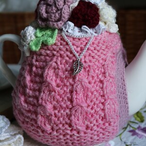 May include: A pink knitted teapot cosy with crocheted flowers and a pearl necklace with a leaf charm. The cosy is on a white tablecloth with embroidered flowers.