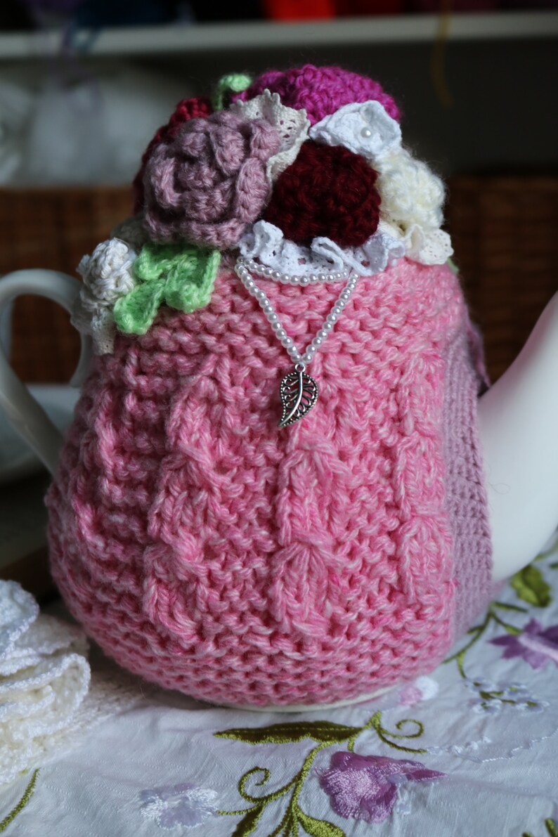 May include: A pink knitted teapot cosy with crocheted flowers and a pearl necklace with a leaf charm. The cosy is on a white tablecloth with embroidered flowers.