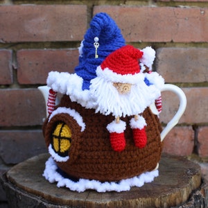 May include: A brown crocheted teapot cozy with a white trim and a window. A gnome wearing a red hat and white beard sits on top of the cozy.