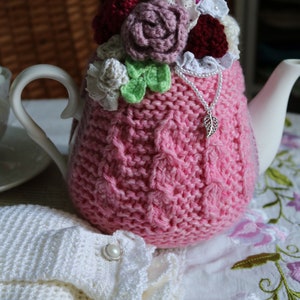 May include: A pink knitted teapot cosy with a crocheted flower design in shades of pink, white, and green. The cosy is adorned with a silver charm and a string of white pearls.