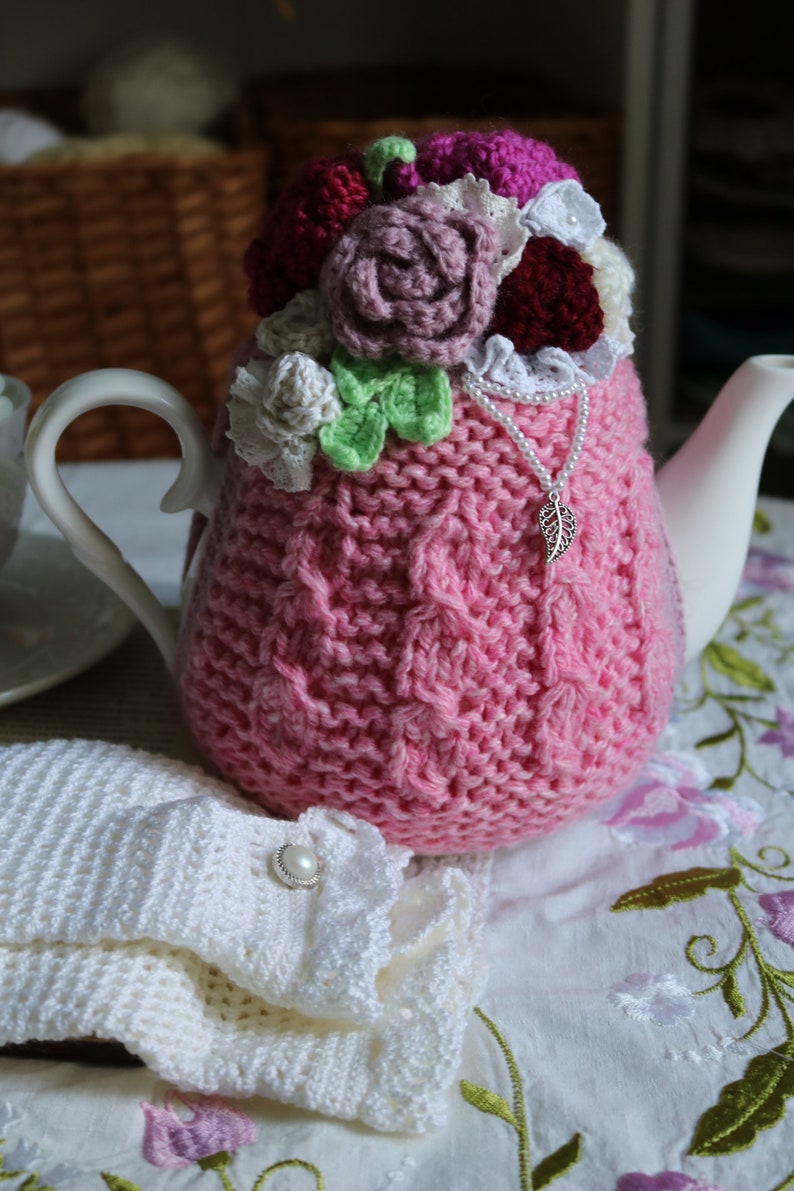 May include: A pink knitted teapot cosy with a crocheted flower design in shades of pink, white, and green. The cosy is adorned with a silver charm and a string of white pearls.