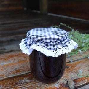 Puede incluir: Un tarro de conserva oscura con una tapa de tela a cuadros blancos y negros, adornada con encaje blanco. El tarro está sobre una superficie de madera, con vegetación en el fondo.