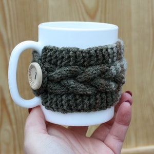 May include: White ceramic mug with a knitted green and brown cozy. The cozy has a wooden button with the word "Handmade" on it.