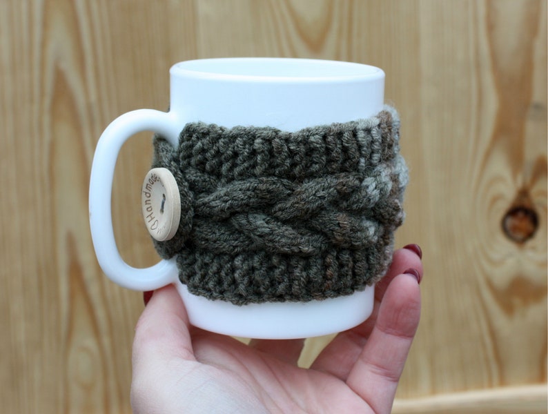 May include: White ceramic mug with a knitted green and brown cozy. The cozy has a wooden button with the word "Handmade" on it.