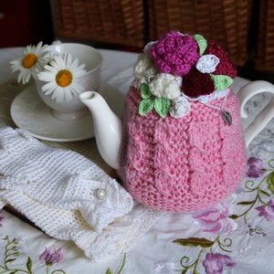 May include: A pink crocheted teapot cosy with a floral design. The cosy is decorated with pink, white and burgundy crocheted flowers. The teapot cosy is sitting on a white tablecloth with embroidered flowers.