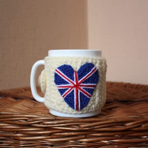Könnte beinhalten: Eine weiße Keramiktasse mit einer gehäkelten Tasse, die ein herzförmiges Union Jack-Design in Rot, Weiß und Blau zeigt.