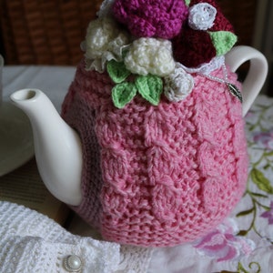 May include: A pink crocheted teapot cosy with a floral design. The cosy features white, pink and burgundy crocheted flowers with green leaves.