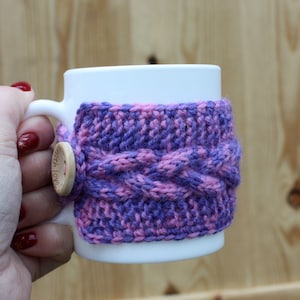 May include: White ceramic mug with a purple and pink knitted cozy. The cozy has a cable knit pattern and a wooden button that says "With Love".