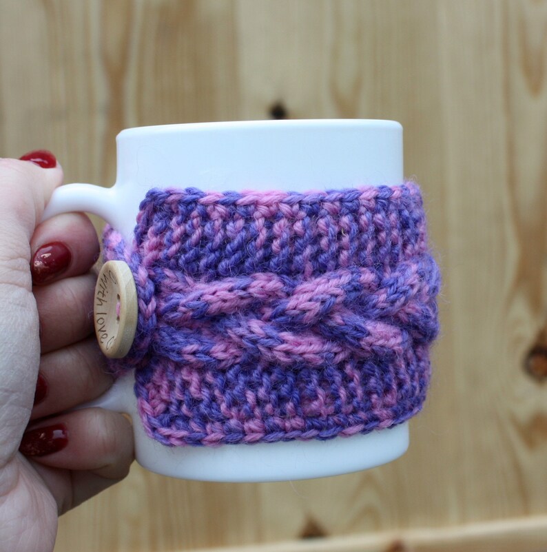 May include: White ceramic mug with a purple and pink knitted cozy. The cozy has a cable knit pattern and a wooden button that says "With Love".