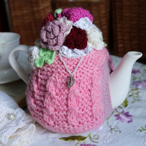 May include: A pink crocheted teapot cosy with a floral design. The cosy features a variety of crocheted flowers in pink, white, green, and burgundy. A silver chain with a leaf charm hangs from the cosy.