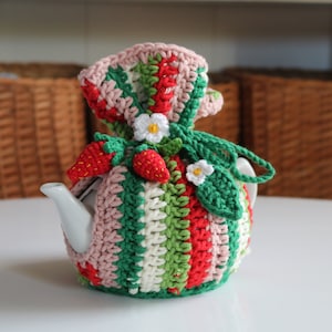 May include: A crocheted teapot cozy with a strawberry and flower design in shades of pink, green, red, and white.