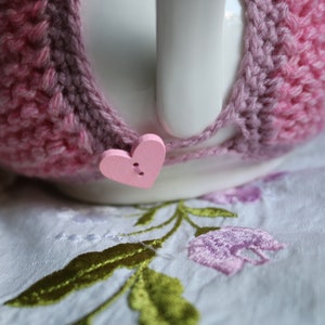 May include: A white ceramic teapot with a pink crocheted cosy. The cosy has a pink heart-shaped button.