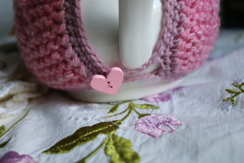 May include: A white ceramic teapot with a pink crocheted cosy. The cosy has a pink heart-shaped button.
