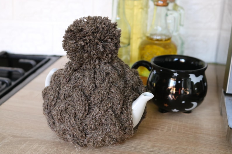 May include: A brown knitted teapot cozy with a fluffy pom pom on top. The cozy is made of a thick, cable knit yarn and is designed to keep a teapot warm.