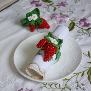 May include: Two crochet strawberry napkin rings with green leaves and white flowers. One ring is wrapped around a white napkin on a white plate. The other ring is on the table. The background is a white tablecloth with floral embroidery.