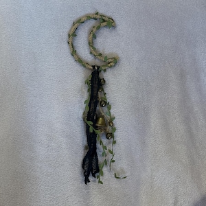 May include: A crescent moon-shaped wall hanging. The moon is made of twisted rope and adorned with small green leaves. A black lace and bell tassel hangs below, with additional small bells and faux greenery.