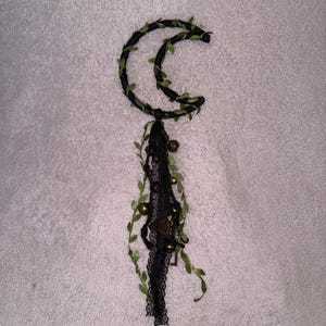 May include: A crescent moon-shaped decoration with black woven material and green leaf accents. The moon is adorned with a black lace and gold-colored beads and charms, creating a whimsical and mystical aesthetic.