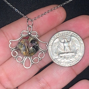 May include: Silver necklace with a delicate filigree pendant featuring a round resin charm filled with dried flowers and foliage. The pendant is about the size of a US quarter.
