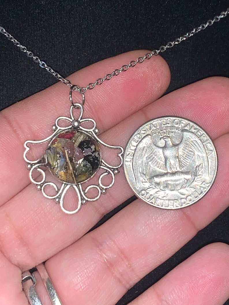 May include: Silver necklace with a delicate filigree pendant featuring a round resin charm filled with dried flowers and foliage. The pendant is about the size of a US quarter.
