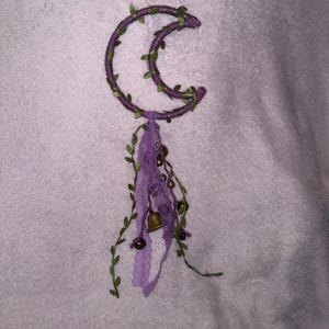 May include: A purple crescent moon-shaped wreath with green leaves and a purple and white tassel with bells hanging from the bottom.