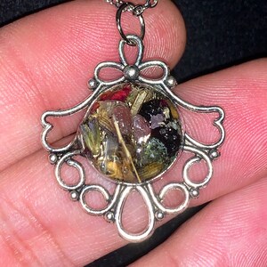 May include: A silver necklace with a round pendant featuring a variety of dried flowers and herbs encased in clear resin. The pendant is surrounded by a decorative silver frame.