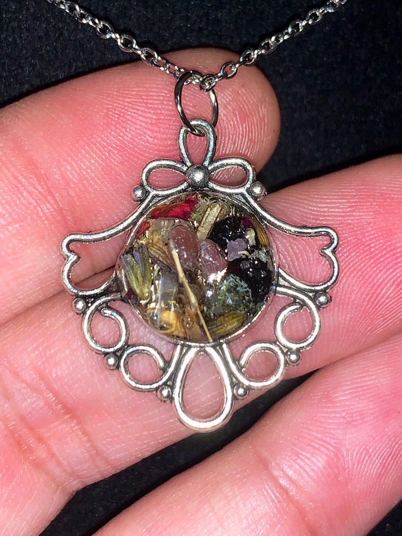 May include: A silver necklace with a round pendant featuring a variety of dried flowers and herbs encased in clear resin. The pendant is surrounded by a decorative silver frame.