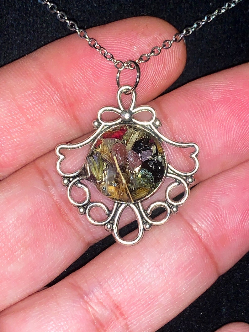 May include: A silver necklace with a delicate filigree pendant. The pendant features a round glass dome filled with dried flowers and herbs.