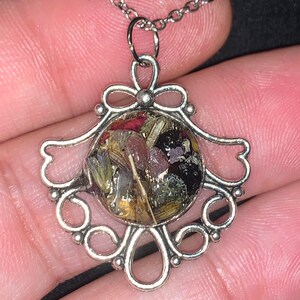 May include: A silver necklace with a delicate, ornate pendant featuring a round, clear resin charm filled with dried flowers and herbs.