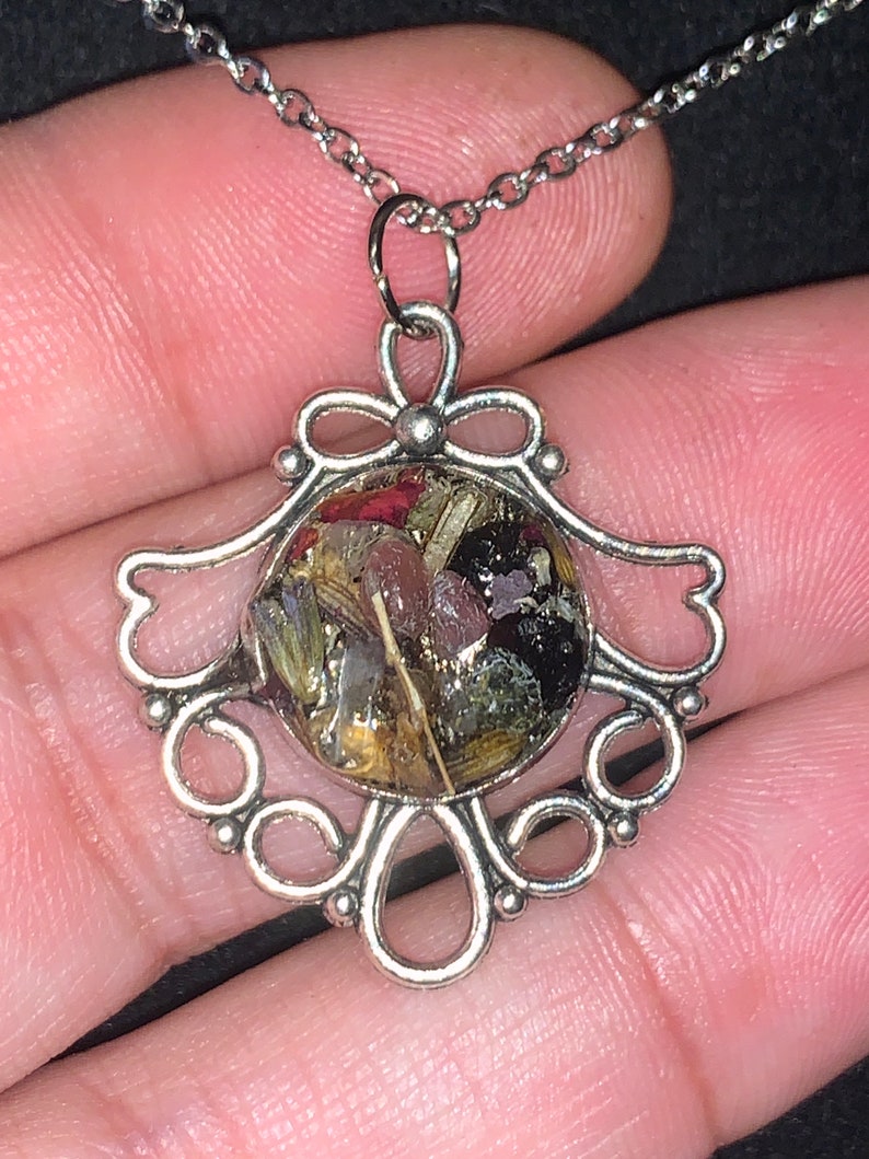 May include: A silver necklace with a delicate, ornate pendant featuring a round, clear resin charm filled with dried flowers and herbs.