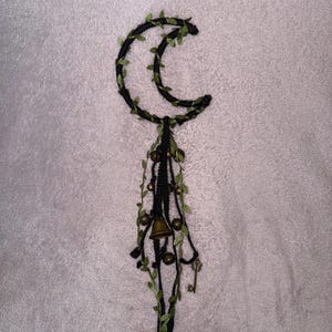 May include: A black crescent moon-shaped wall hanging with green leaf accents. The moon is adorned with hanging strands, small bells, and a tiny key. The design is set against a textured, off-white background.