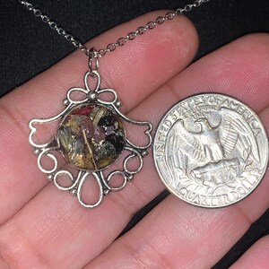 May include: A silver necklace with a pendant featuring a round, clear resin charm filled with dried flowers and herbs. The pendant is attached to a silver chain and is about the size of a quarter.