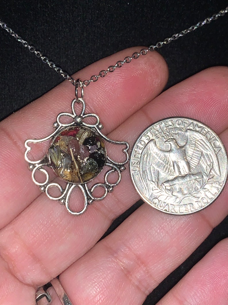 May include: A silver necklace with a pendant featuring a round, clear resin charm filled with dried flowers and herbs. The pendant is attached to a silver chain and is about the size of a quarter.