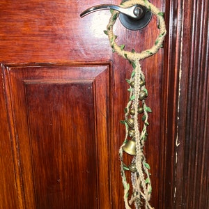May include: A brown wooden door with a silver door handle and a handmade door knocker. The knocker is made of twine and has a small bell attached to it. The knocker is decorated with green leaves.