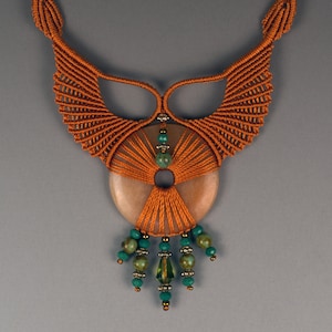 以下が含まれることがあります： 茶色のマクラメネックレスに、大きな丸い薄い茶色の石のペンダントが付いています。ペンダントの中央には穴があり、その周りを小さな緑色のビーズが囲んでいます。ネックレスは太い編組コードで作られています。