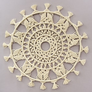 May include: A large, circular macrame wall hanging with a cream color. The macrame has a detailed design with multiple layers of knots and fringe around the edges.