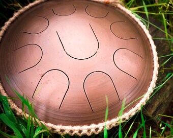 Könnte beinhalten: Ein braunes Stahl-Handpan mit einer Kordelkante liegt auf einem Bett aus grünem Gras. Das Handpan hat eine runde Form und ist mit einem Muster aus kleinen, geschwungenen Linien verziert.