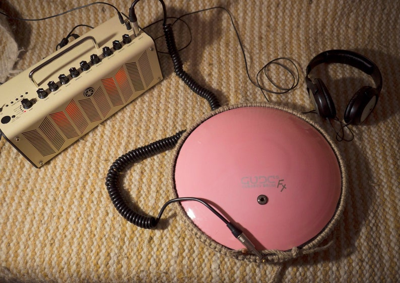 May include: A pink round drum with a rope border and the text "AUDIO FX" on the surface. A black coiled cable connects the drum to a beige guitar amplifier with a black handle. A pair of black headphones are on the right side of the image.