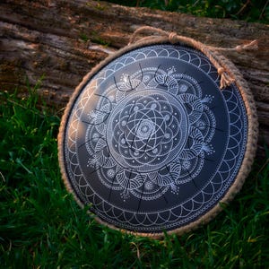 May include: A black handpan with a white mandala design. The handpan is resting on a log surrounded by green grass.