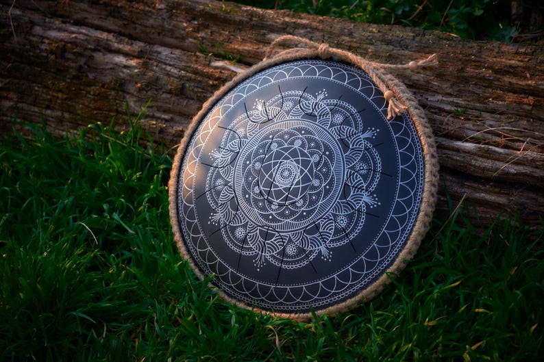 May include: A black handpan with a white mandala design. The handpan is resting on a log surrounded by green grass.