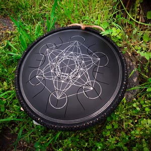 May include: A black steel handpan with a geometric design, featuring a circular frame with a rope handle. The handpan is resting on a bed of green grass.
