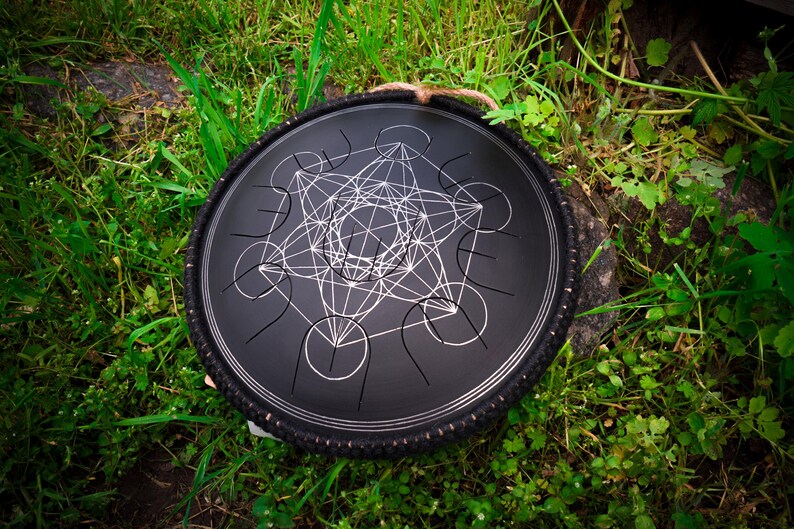 May include: A black steel handpan with a geometric design, featuring a circular frame with a rope handle. The handpan is resting on a bed of green grass.