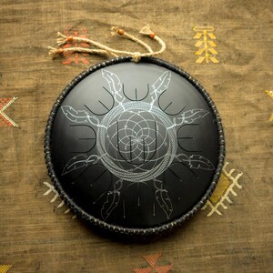 May include: A black handpan with a silver geometric design. The handpan is on a brown fabric background with white and yellow patterns.