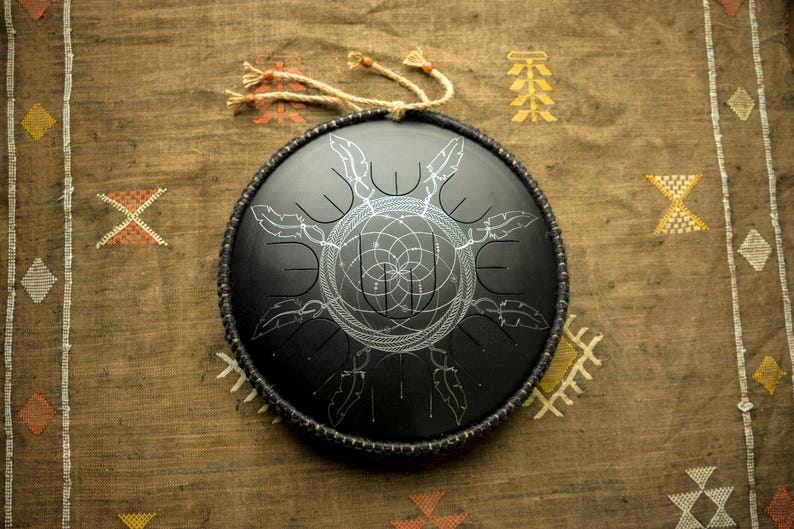 May include: A black handpan with a silver geometric design. The handpan is on a brown fabric background with white and yellow patterns.