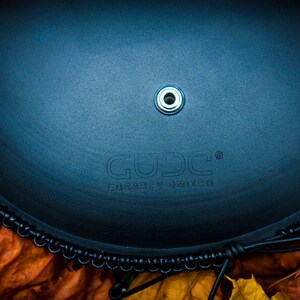 May include: A black leather drum with a black metal rim and a silver metal ring in the center. The drum has the word "GUDJ" and the text "FRABEN INGUIR" printed on it.